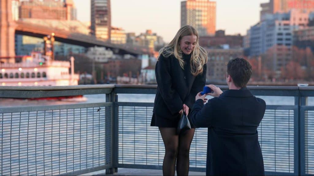 A man kneeling and proposing to a woman with a ring by a riverside city view.