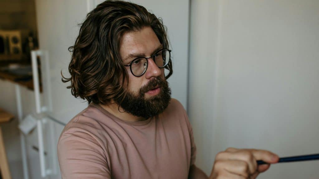 A man with long wavy hair and glasses holds a blue pen while looking to the side.