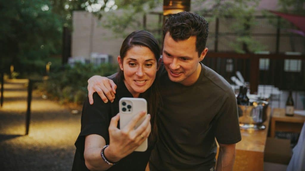 A smiling woman takes a selfie with a man who has his arm around her.