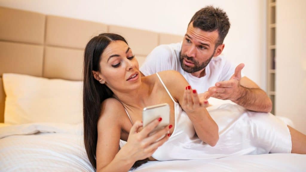 A woman lies on a bed looking at a phone while a man gestures behind her.