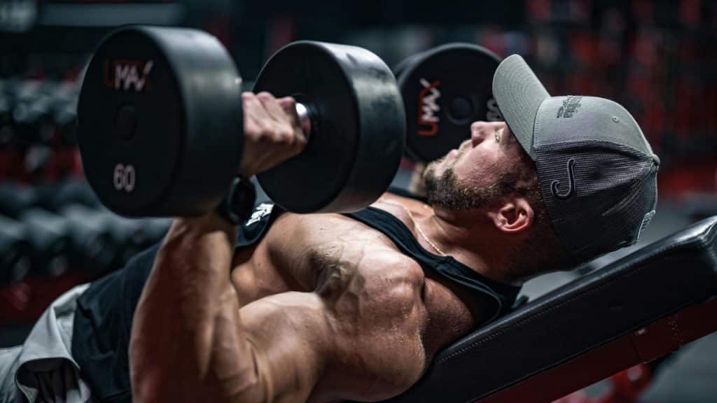 A man in a grey cap performs a chest press with heavy dumbbells in a gym.