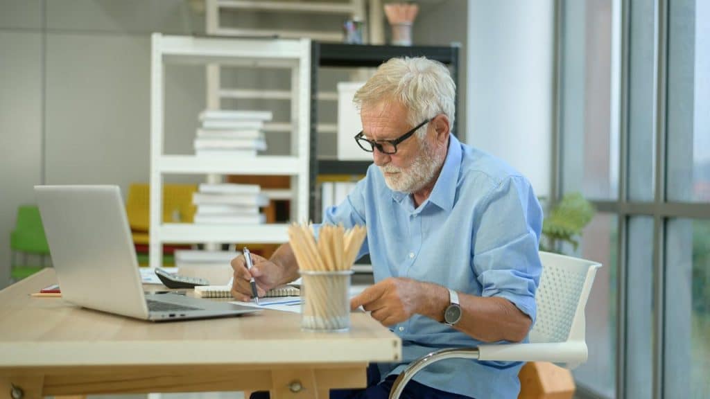An older man with glasses writes in a notebook at a desk with a laptop.
