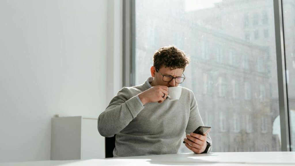 A man wearing glasses sitting by a window, drinking coffee while looking at his phone.