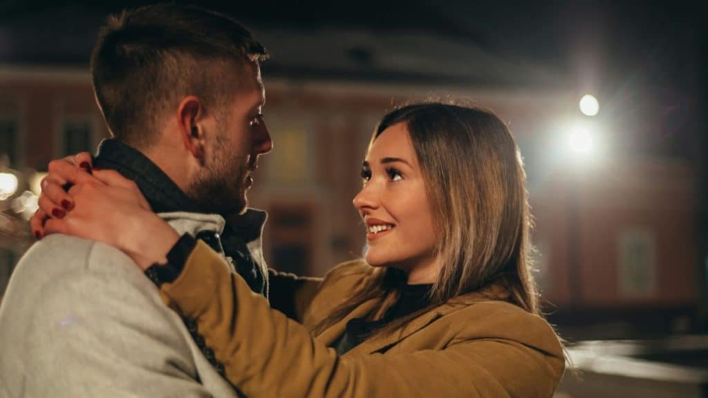 A man and a woman embrace outdoors at night, looking at each other with smiles.
