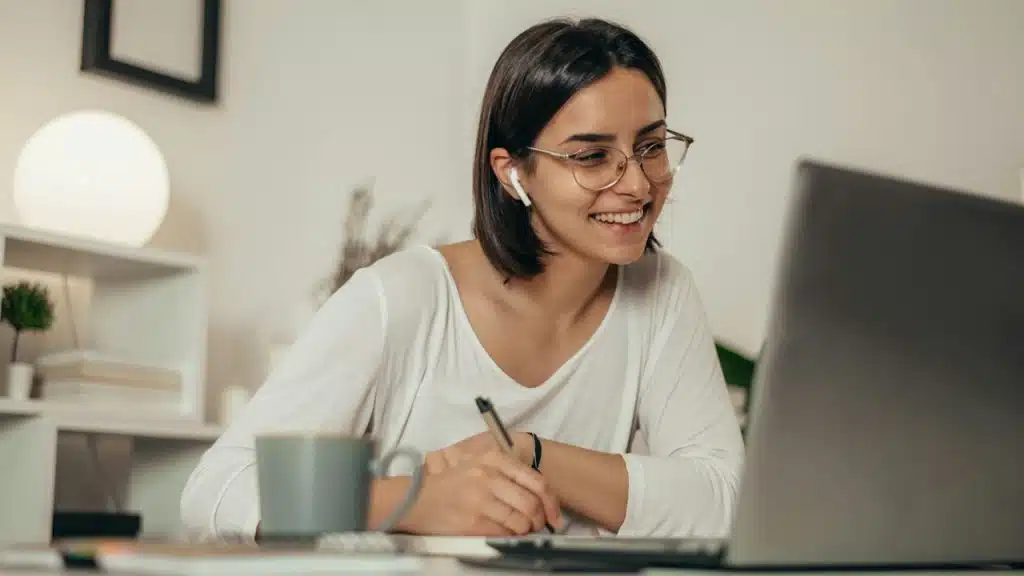 A smiling woman who is wearing glasses is writing on a paper while looking at a laptop screen.
