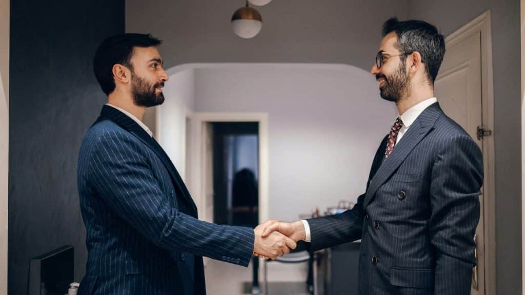 Two men in dark pinstriped suits shake hands while standing inside an office hallway.