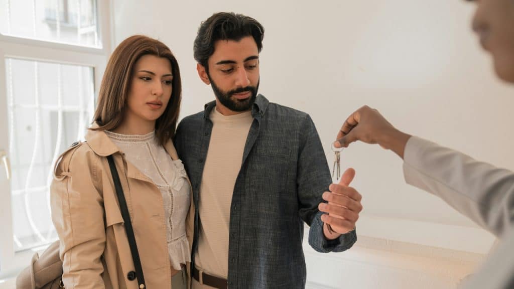 A person hands keys to a couple standing in a bright indoor room.