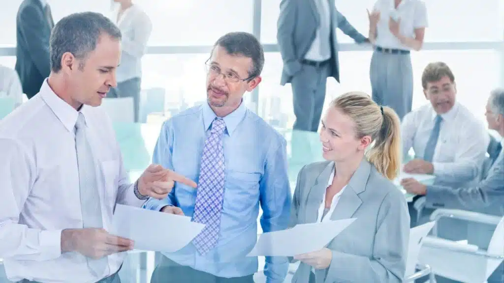 Three professionals stand in an office, two holding papers and talking to each other.