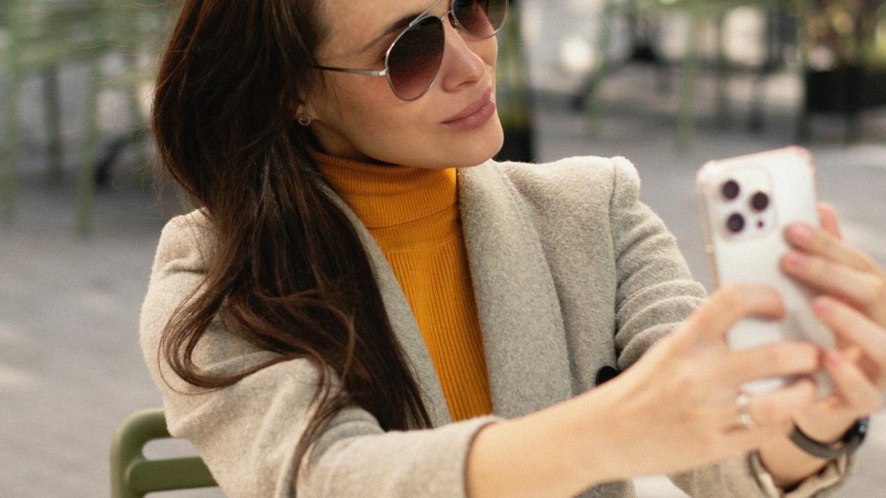 A woman in sunglasses taking a selfie outdoors with her smartphone.