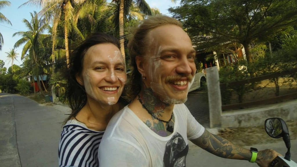 A smiling man and woman with white paste on their faces ride a scooter outdoors.
