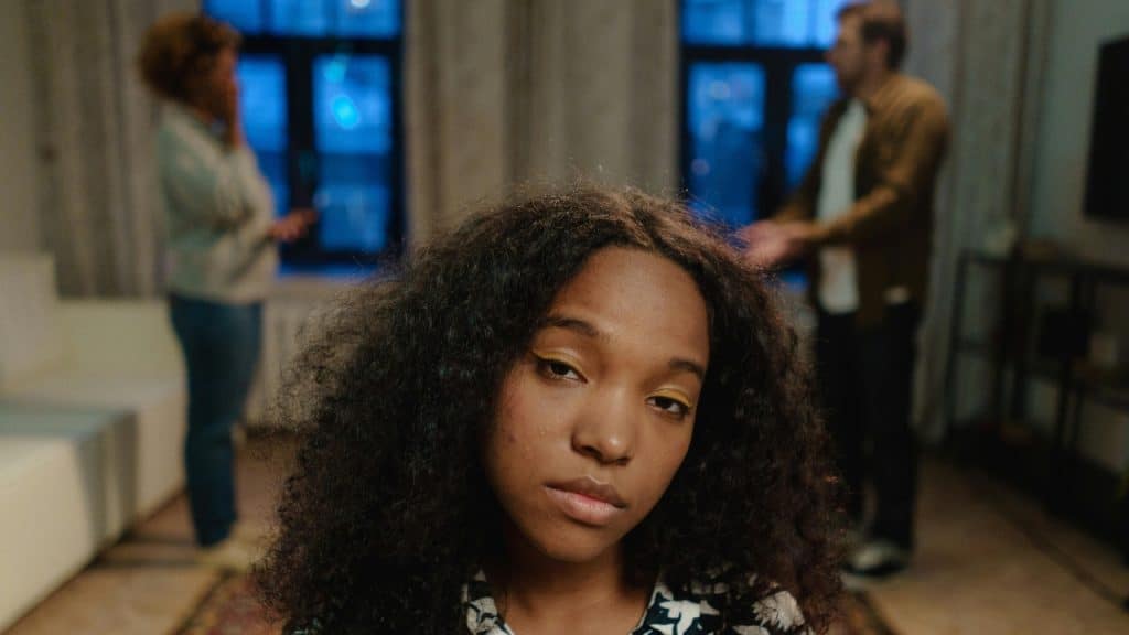 A young woman with curly hair looks at the camera while parents argue in background.