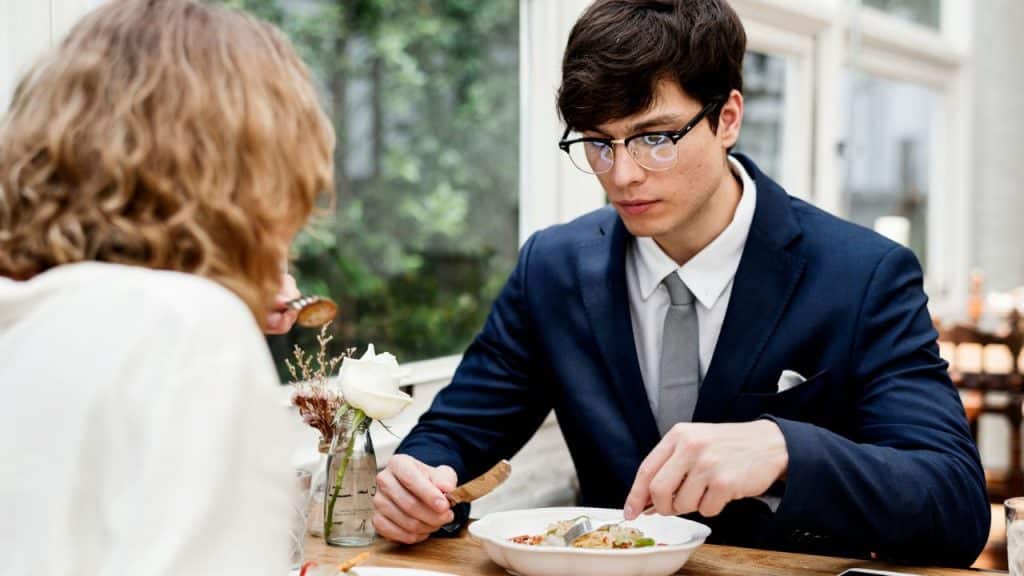 A man in a suit looks down at his plate while eating dinner across from someone.