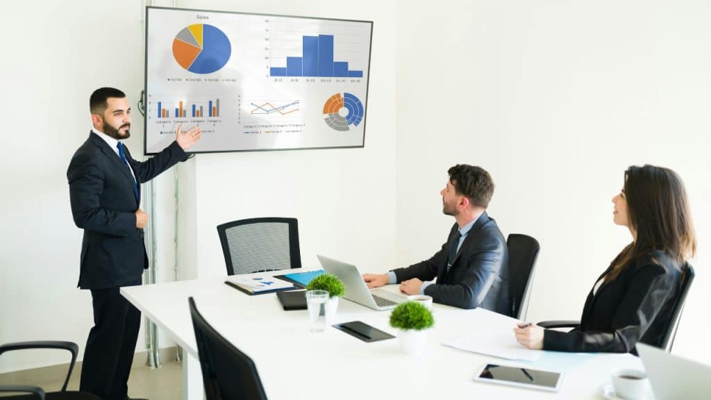 A man in a suit gives a presentation with charts to two colleagues.