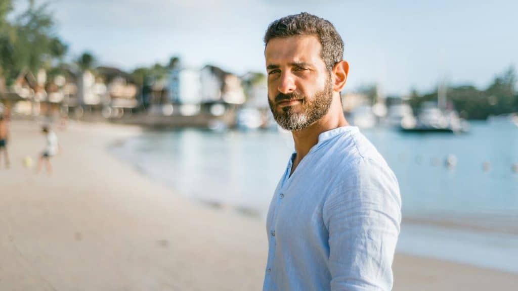 A bearded mature man looking at the camera as he stands on a beach.