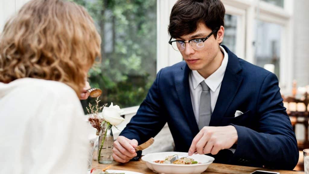 A man in a suit and glasses eats while seated across from a woman.
