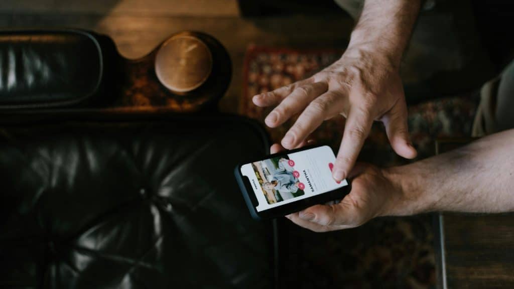 Hands holding a smartphone display a dating profile with a heart and X icon.