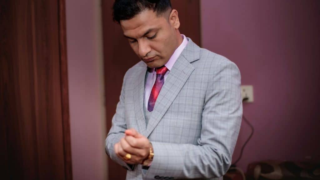 A man in a light gray plaid suit adjusts his sleeve while looking down.