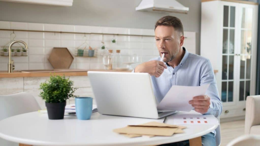 A man in a blue shirt sits at a table using a laptop and reviewing documents.