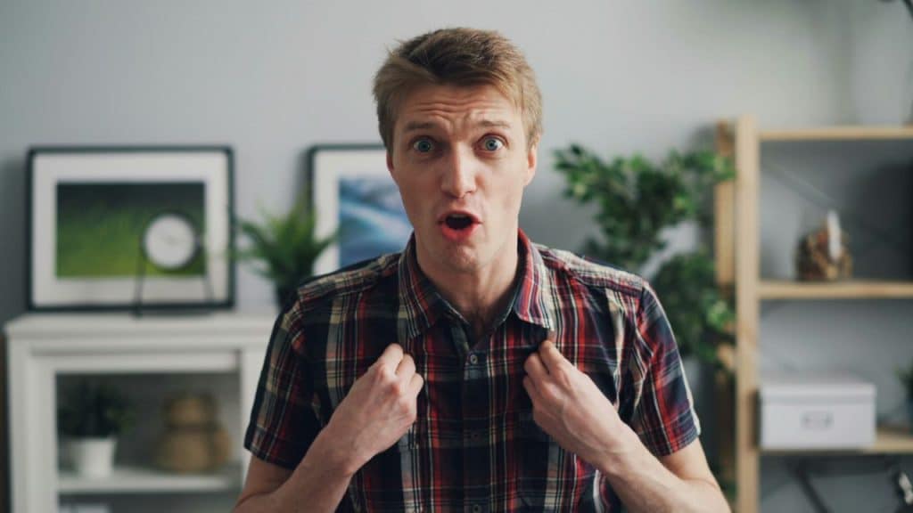 A man with an open-mouthed expression holds the collar of his plaid shirt indoors