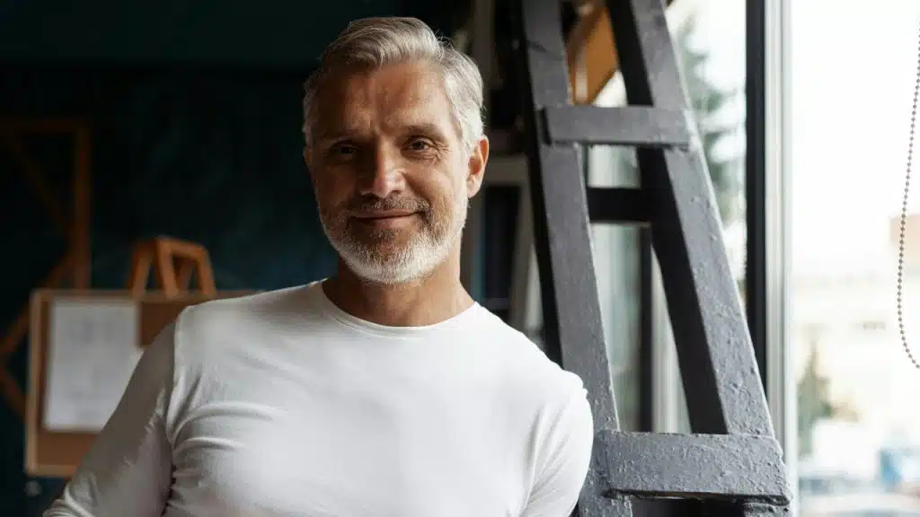 A smiling, gray-haired man in a white shirt stands next to a black ladder.