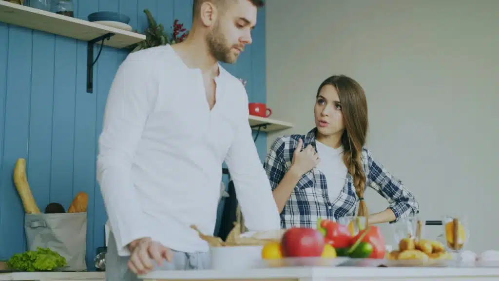 A woman gestures while talking to a man standing in a kitchen with vegetables.