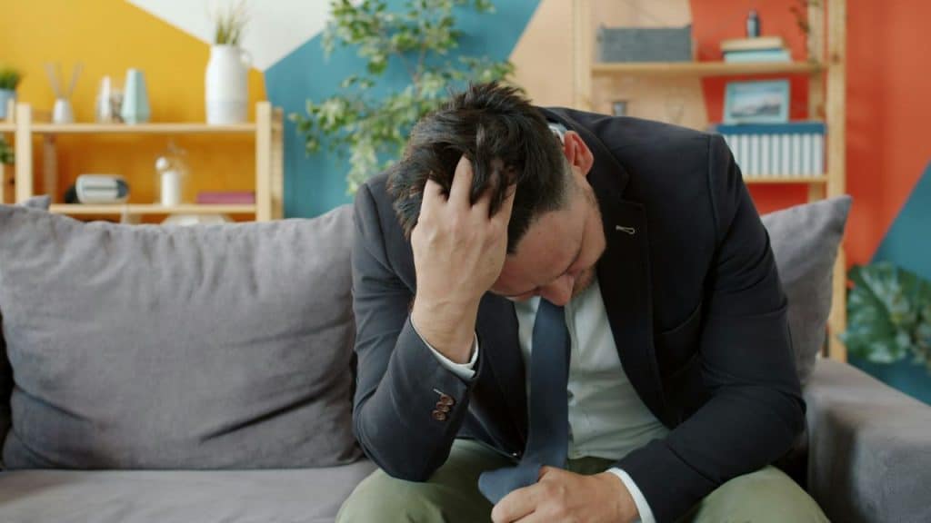 A man in a suit jacket sits on a sofa holding his head.