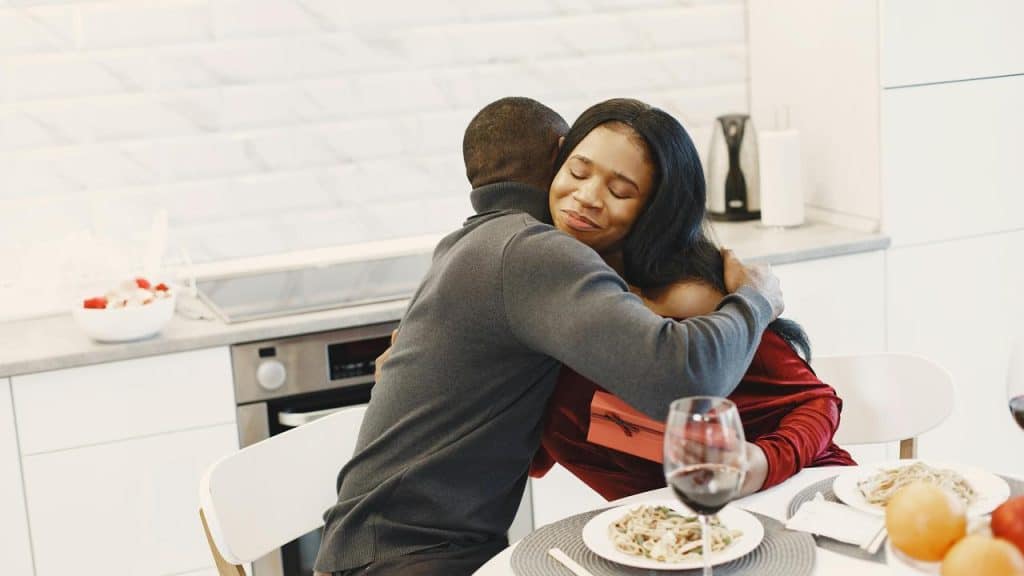 A couple embracing at a dinner table with wine and a gift in a bright kitchen.