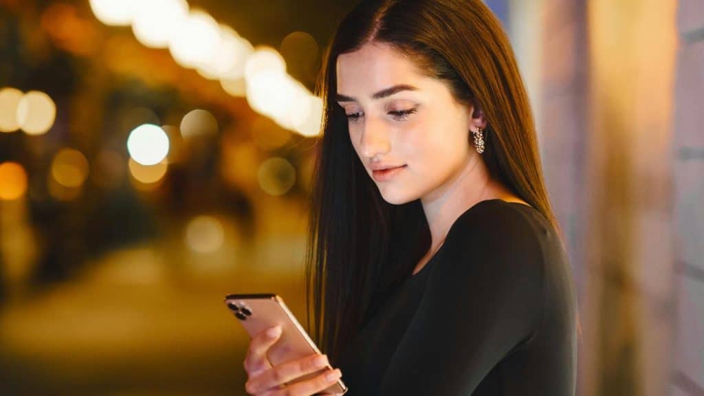 A woman looking at her smartphone at night with blurred city lights behind her.