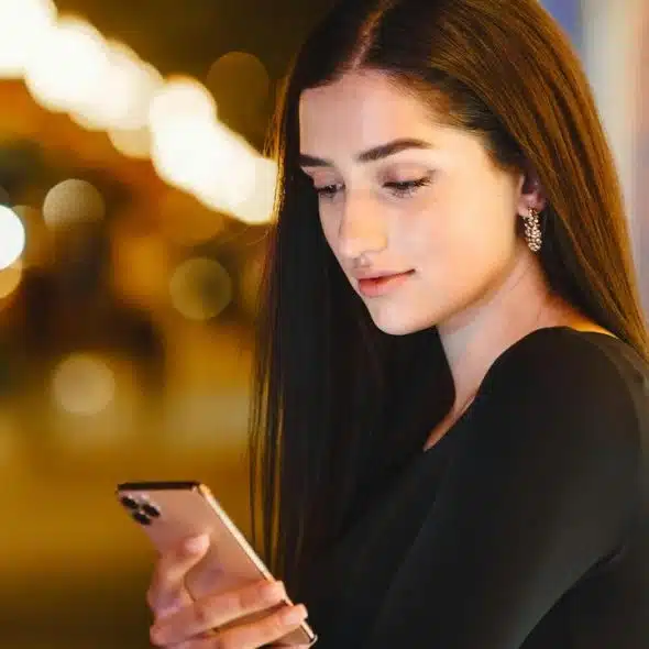 A woman looking at her smartphone at night with blurred city lights behind her.