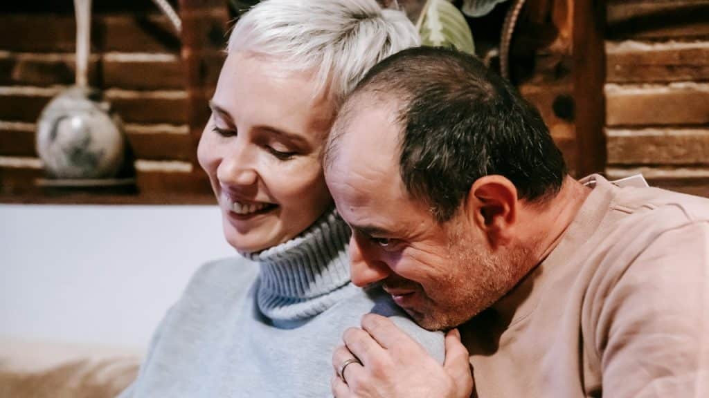 A woman with short blonde hair smiles while a man rests his head on her.