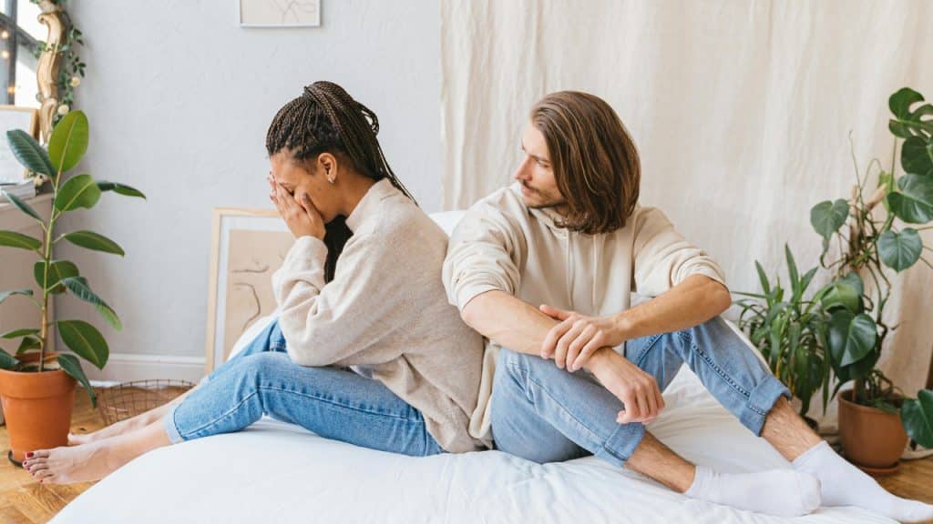 A woman covers her face while sitting back-to-back with a man on a bed.