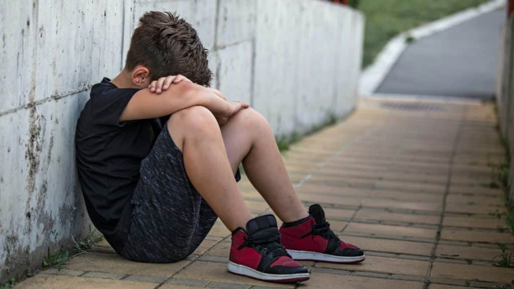 A young boy sits on the ground against a concrete wall, head on knees.