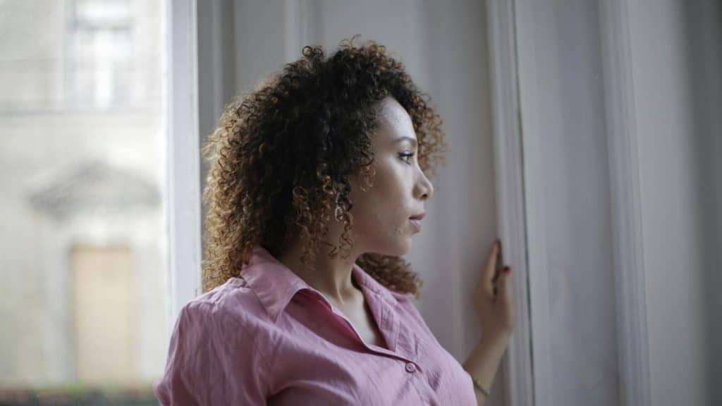 A woman in a pink shirt looking out of a window thoughtfully.