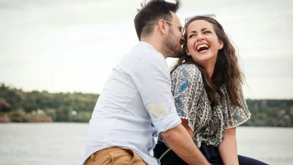 A man whispers into the ear of a woman who is laughing outdoors near water.