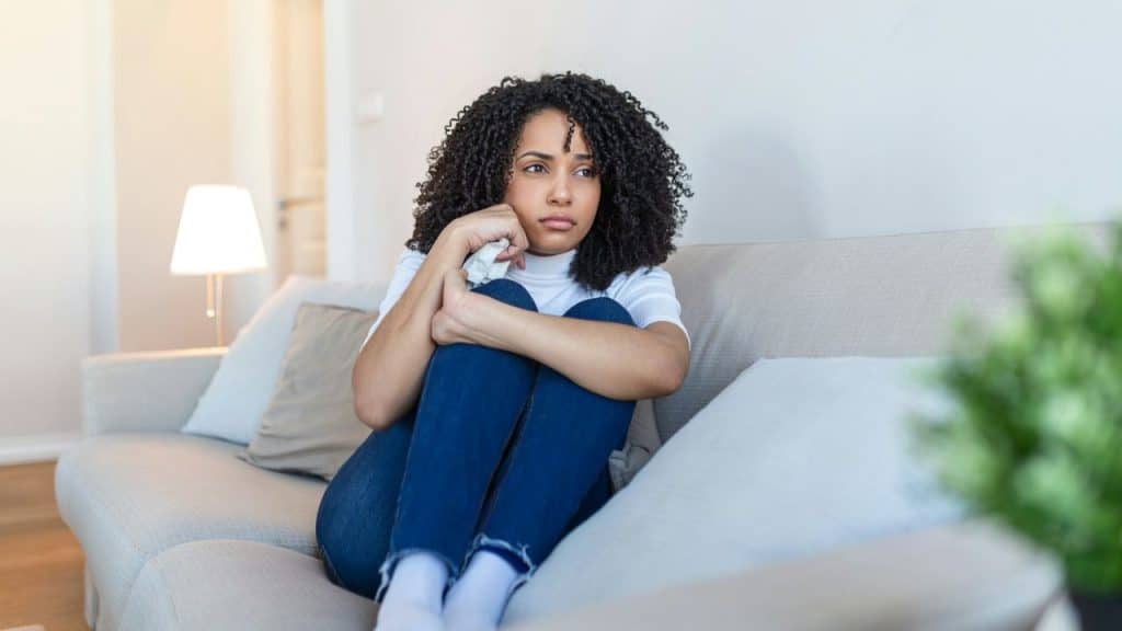 A woman with curly hair sits on a couch, hugging her knees and looking away.