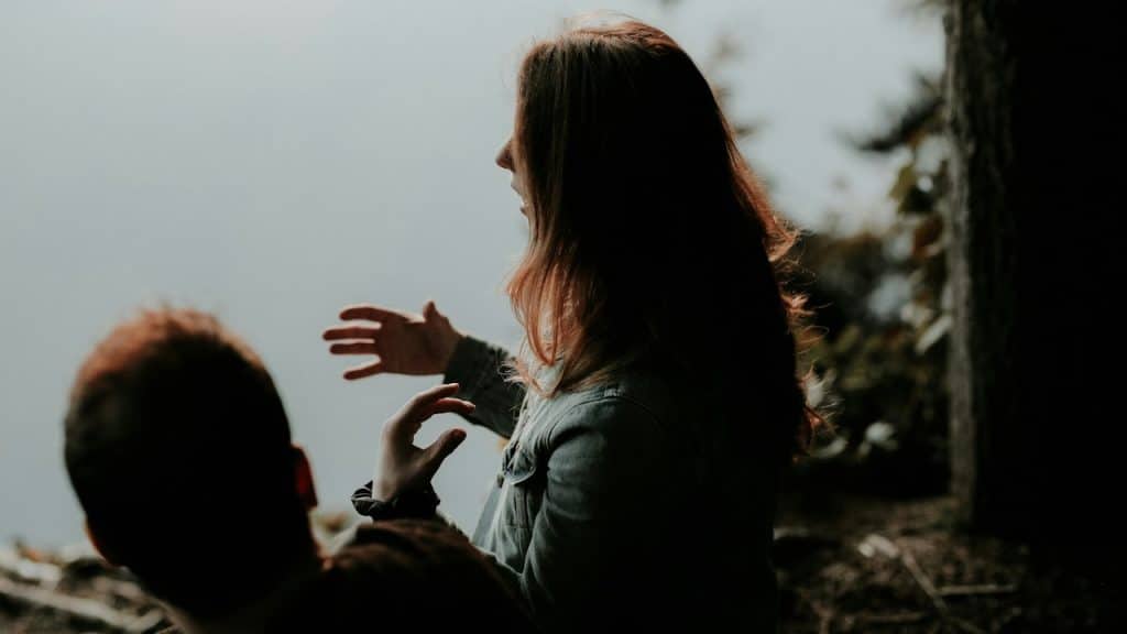 A woman in a denim jacket gestures with open arms while talking to a man outdoors.
