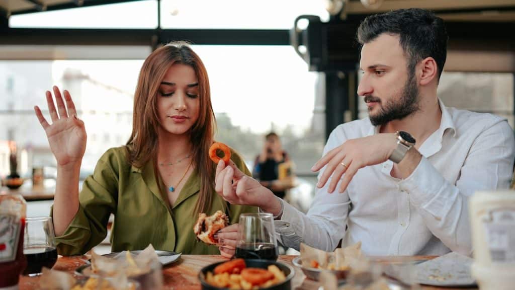 A woman refusing food offered by a man while sitting together at a restaurant table.