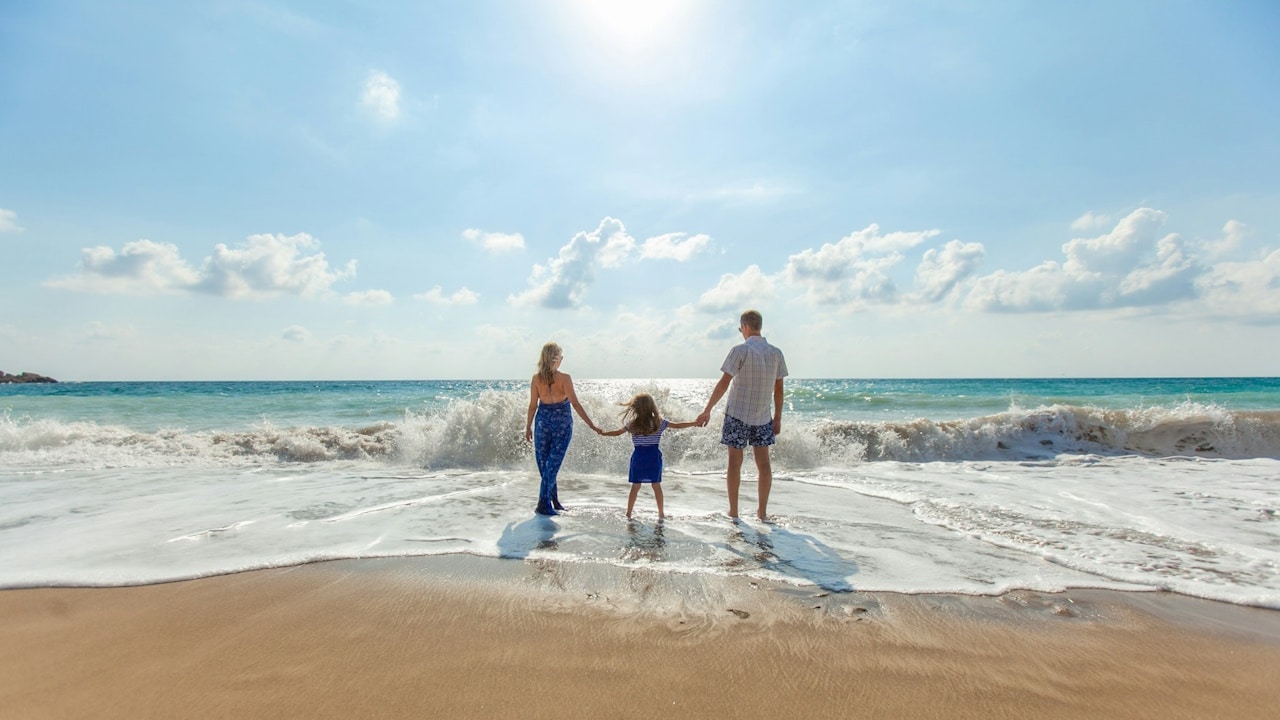 A couple standing with their kid at the beach and holding hands in front of a small wave.