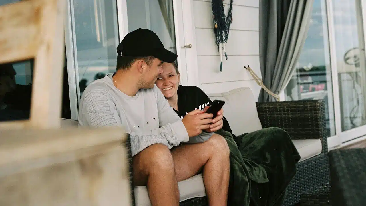 A smiling woman wearing a blanket is leaning against a man wearing a cap and using his mobile while they both sit on a couch outside their home.
