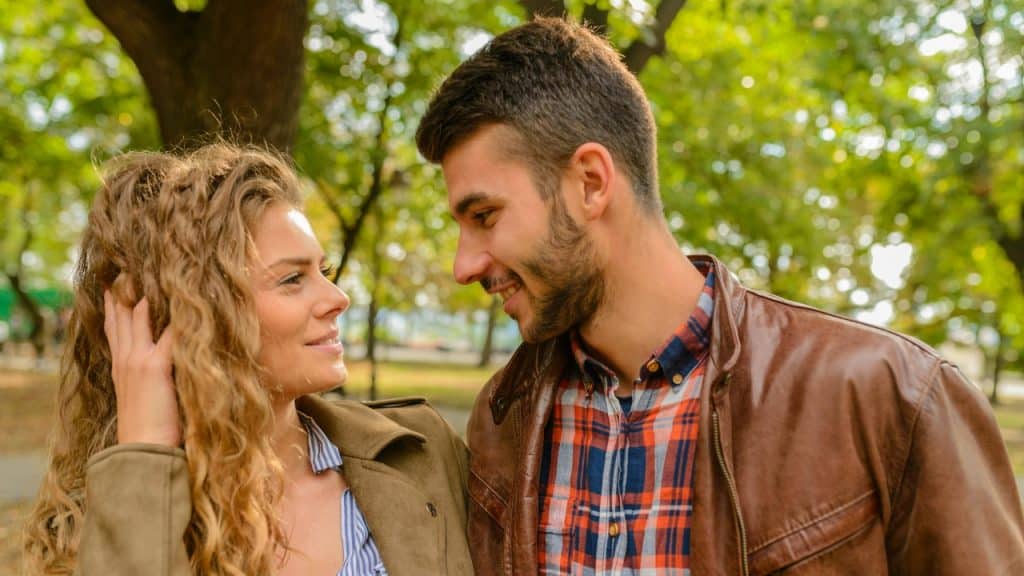 A man and woman look at each other and smile outdoors with green trees in background.