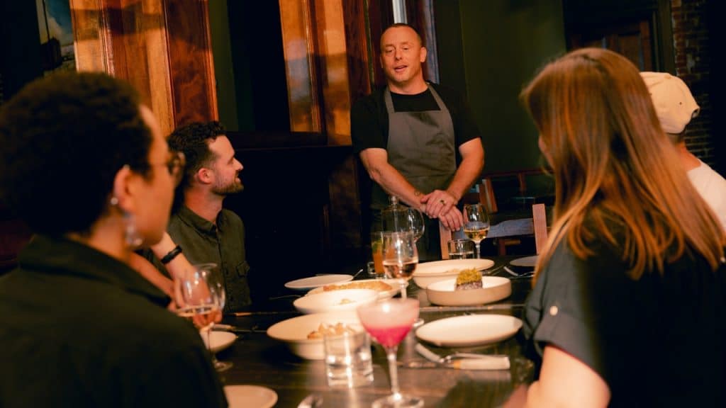 A man in an apron stands at the head of a table with seated people.