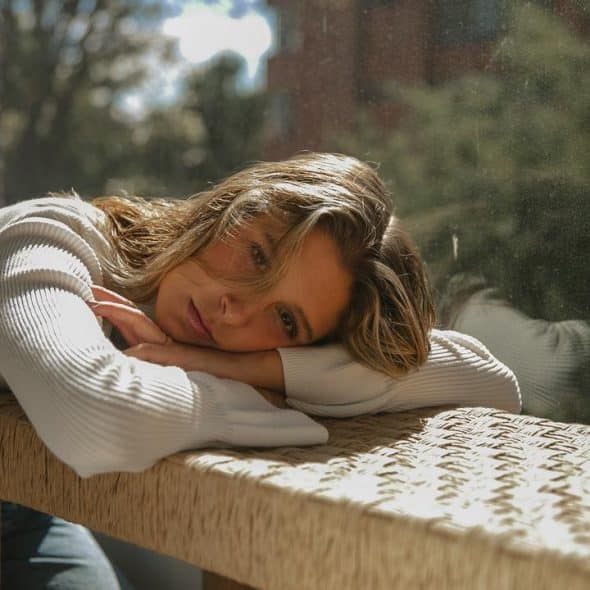 A woman resting her head on a woven bench near a sunlit window.
