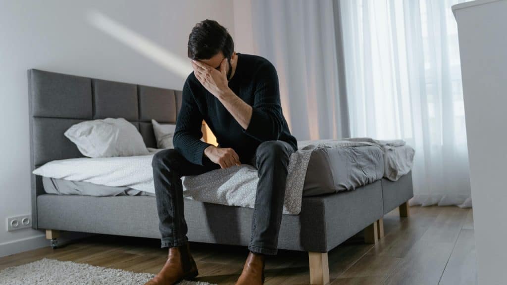 A man in a dark sweater sits on a bed, covering his face.