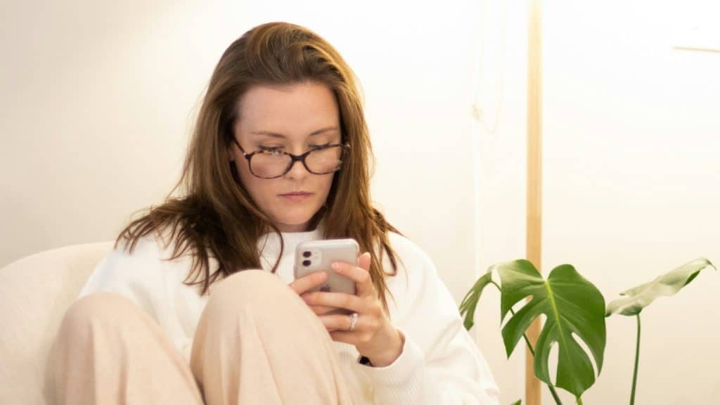 A woman with glasses sits in a chair, looking down at her smartphone.