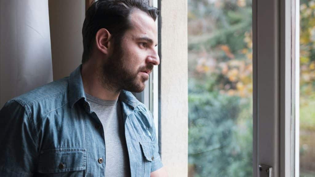 A man with a beard and denim shirt gazes out of a window with a serious expression.