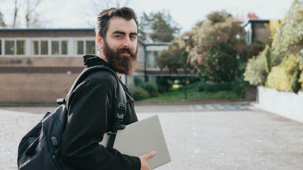 A man holding a laptop and a backpack is looking at the camera.