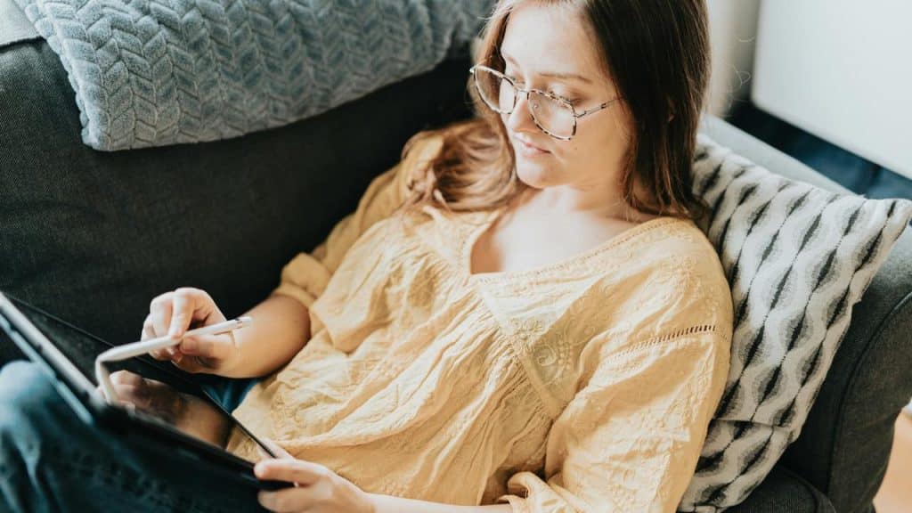 A woman wearing glasses using a tablet while sitting on a couch.
