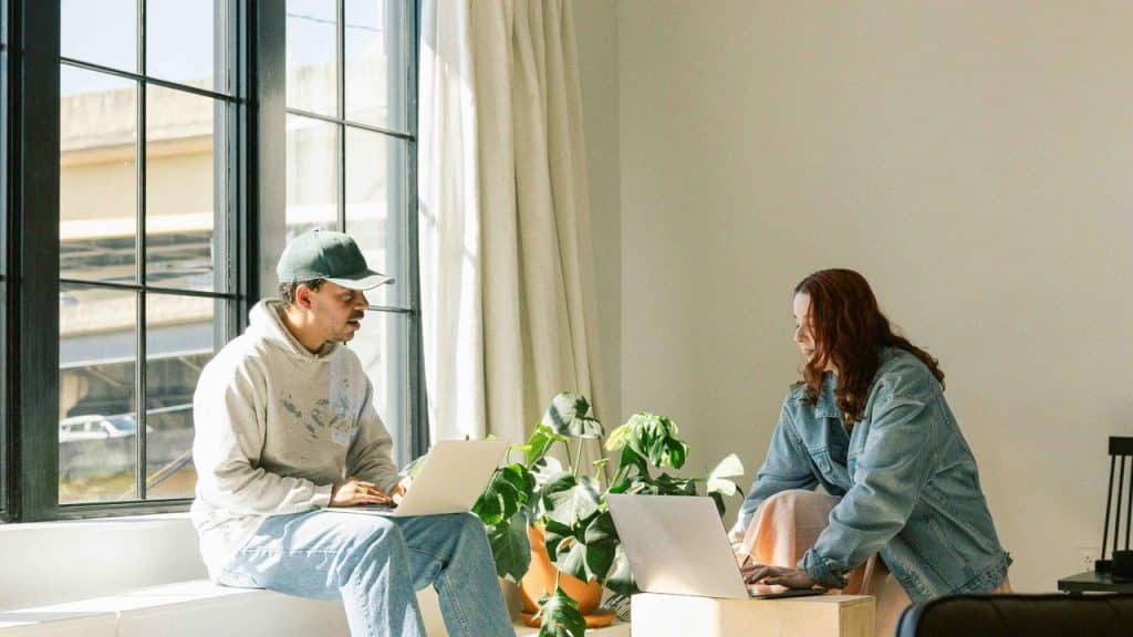 A man and woman working on laptops near a large window.
