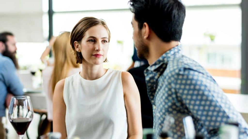 A woman in a white sleeveless top looks toward a man in a patterned shirt.