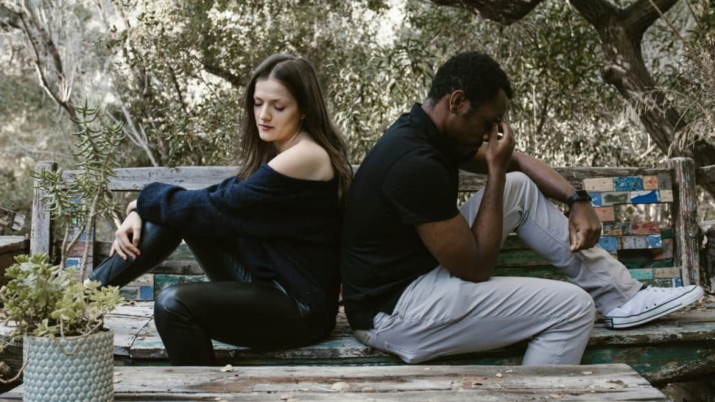 A woman and man sit back-to-back on a wooden bench outdoors looking downward.